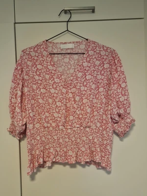 Blommig rosa blus med volang - Söt blommig blus i rosa och vitt med v-ringning och volangkant nedtill. Blusen har trekvartsärm med resår vid ärmslut och smockad midja för en feminin siluett. Perfekt till jeans eller kjol för en trendig look.