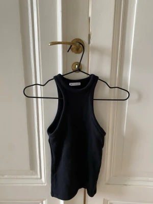 Svart ribbad linnetopp - Snygg svart ribbad linnetopp med hög halsringning och ärmlös design. Perfekt att styla till jeans eller kjol för en clean och enkel look. Materialet känns mjukt och stretchigt.