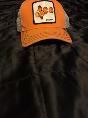 Orange truckerkeps med clownfisk - Snygg orange truckerkeps med vit mesh baktill och ett broderat märke framtill med en clownfisk och texten 'CLOWN'. Kepsen har böjd skärm och justerbar snapback. Perfekt för dig som gillar färg och coola detaljer.