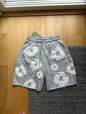 Grå denim tears shorts - Grå shorts med elastisk midja och snörning, dekorerade med stora vita molnliknande tryck över hela plagget. Mjukt material som känns skönt mot huden och avslappnad passform. Perfekta för chill dagar eller när du vill sticka ut lite extra.