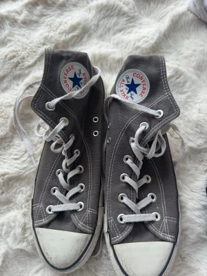 Grå Converse All Star High Tops - Klassiska grå Converse All Star high top sneakers i canvas med vita gummisulor och tåhättor. Skorna har vita snören och den ikoniska loggan på sidan. Perfekta för en avslappnad och cool stil.
