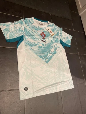 Portugal tränings t-shirt Puma - Snygg Portugal tränings t-shirt från Puma i vitt och turkost med vågigt mönster. Har Portugal-emblemet på bröstet och Puma-logga. Tillverkad i lätt och ventilerande material, perfekt för träning eller sport.