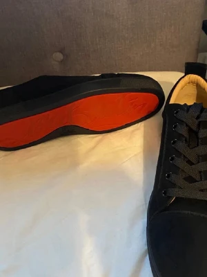 Louboutins - Snygga svarta sneakers från Christian Louboutin med klassisk röd sula. Skorna har en låg profil, svarta skosnören och ovandel i mocka. Perfekta för dig som vill sticka ut med en ikonisk detalj på fötterna.