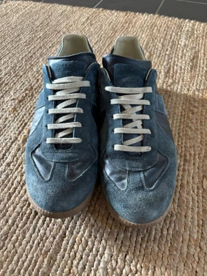 Blå gats från Maison Margiela - Snygga blå sneakers från Maison Margiela med ovandel i mocka och läderdetaljer. Skorna har vita snören och en klassisk gummisula med mönster undertill. Perfekta för dig som gillar stilrena och unika sneakers.