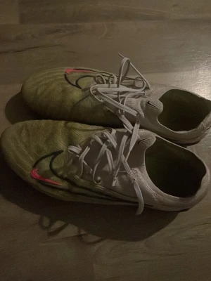 Vita och gröna Nike fotbollsskor - Säljer ett par vita och gröna fotbollsskor från Nike med klassisk snörning och en svart samt röd swoosh på sidan. Skorna har låg profil och är designade för bra bollkontroll på planen. Perfekta för dig som vill ha stilrena och funktionella skor till match eller träning.
