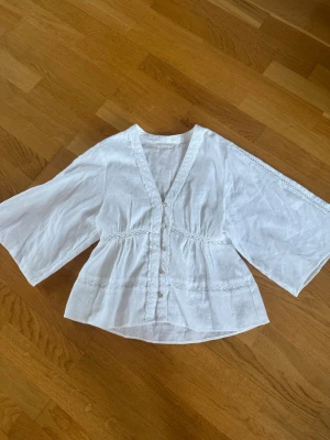 Vit blus med vida ärmar och spetsdetaljer - Söt vit blus med v-ringning, knappar framtill och vida trekvartsärmar. Blusen har fina spetsdetaljer och lätt rynkad midja för en feminin siluett. Perfekt till jeans eller kjol för en trendig look.