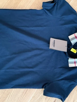 Marinblå Burberry skjorta  - Pris diskuteras vid snabb affär. Snygg marinblå skjorta från Burberry med klassisk rutig krage i beige, svart och rött. Skjortan har korta ärmar och knappar vid halsen. Tillverkad i bomull för en skön känsla och stilren look.