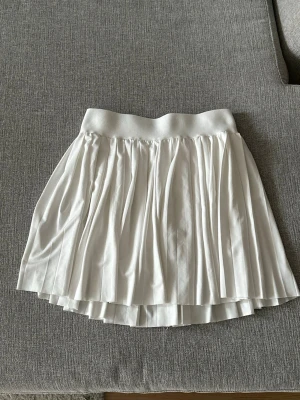 Vit plisserad kortkjol med shorts - Supersöt vit plisserad kortkjol med inbyggda shorts under för extra komfort. Kjolen har bred resår i midjan och ett flowy fall som ger en sportig och fräsch look. Aldrig använd, endast testad!