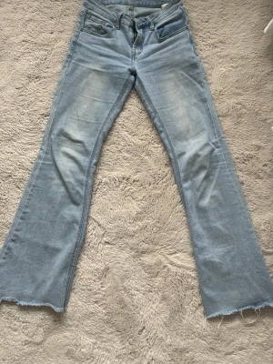 Ljusblå bootcut jeans med rå kant - Säljer ett par ljusblå bootcut jeans med slitningar och råa, avklippta benslut. Jeansen har klassisk femficksmodell och en skön, lite stretchig denim. Perfekta för en avslappnad och trendig look.