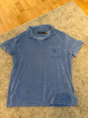 Blå frotté t-shirt från Polo Ralph Lauren - Blå t-shirt i mjuk frotté från Polo Ralph Lauren. Klassisk krage, korta ärmar och bröstficka med broderad logga. Perfekt för en avslappnad och cool stil.