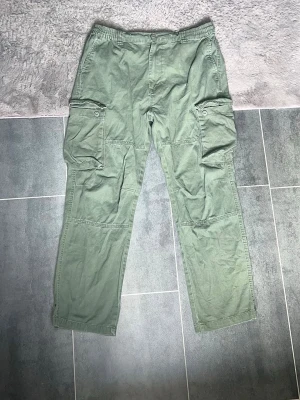 Gröna/gråa 2 cargobyxor  - Snygga gröna/gråa cargobyxor med raka ben och stora sidofickor med lock och knapp. Byxorna har elastisk midja och bakfickor med knapp. Perfekta för en avslappnad och trendig look. Tillverkade i ett mjukt bomullstyg.