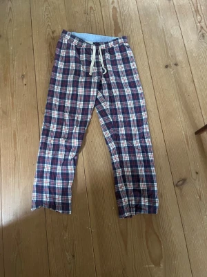 Rutiga pyjamasbyxor med dragsko - Säljer ett par rutiga pyjamasbyxor i rött, blått och vitt med vit dragsko i midjan. Byxorna har avslappnad passform och är perfekta för mysiga hemmakvällar. Tillverkade i mjukt bomullstyg för extra komfort.