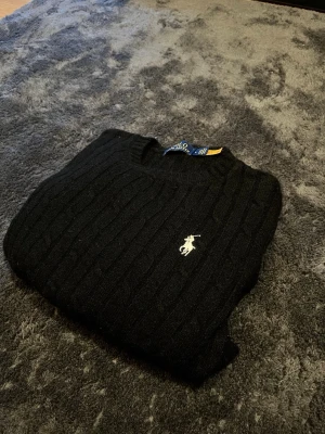 Svart kabelstickad tröja Polo Ralph Lauren - Kabelstickad svart tröja från Polo Ralph Lauren med klassisk rund halsringning och den ikoniska broderade loggan i vitt på bröstet. Tröjan har långa ärmar och ribbade muddar vid ärmslut och nederkant. Perfekt för en stilren och tidlös look. Tröjan är krympt från första tvätten tyvärr och passar därefter som en S. (Tvättade i för hög värme). Tröjan kom därför aldrig till användning. 