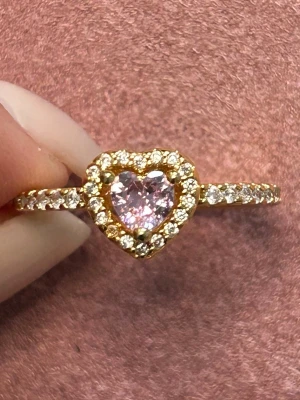 Guldring med hjärtformad rosa sten - Supersöt guldfärgad ring med en hjärtformad rosa sten i mitten, omgiven av gnistrande små klara stenar runt hela hjärtat och längs bandet. Perfekt för dig som gillar romantiska och glittriga accessoarer. Ringen har en elegant och trendig design.