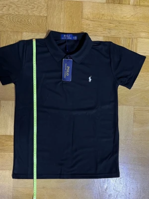 Svart pikétröja från Polo Ralph Lauren - Klassisk svart pikétröja från Polo Ralph Lauren med vit broderad logga på bröstet. Tröjan har krage, korta ärmar och knappar vid halsen. Perfekt till jeans eller shorts för en clean och stilren look.