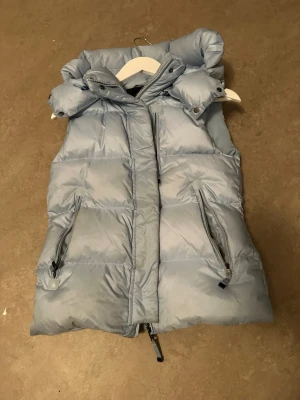 Ljusblå dunväst från Cedrico - Snygg ljusblå dunväst från Cedrico med hög krage och avtagbar huva. Västen har två dragkedjeförsedda fickor och quiltad design. Perfekt för lager på lager och ger en cool look till din outfit.