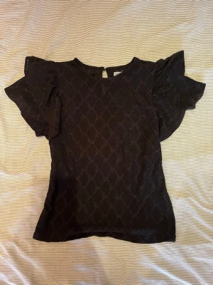 Blus med volangärmar - Snygg svart blus med volangärmar och spets-aktigt mönster. Blusen har rund halsringning och knapp i nacken. 