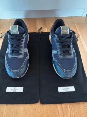 Blå sneakers från Valentino - Snygga sneakers från Valentino i marinblått tyg och mocka med detaljer i grått och mörkblått. Skorna har svarta skosnören, vit sula och klassisk låg silhuett. Perfekta för dig som vill ha en stilren och exklusiv look.