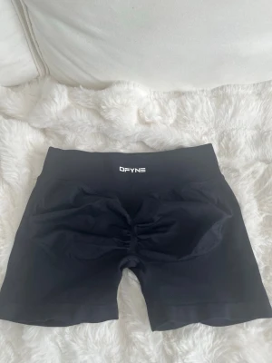 Svarta träningstights shorts från DFYNE - Säljer ett par svarta träningstights shorts från DFYNE med hög midja och scrunch-detalj bak. De är tillverkade i ett stretchigt syntetmaterial som sitter tight och bekvämt. Perfekta för gymmet eller andra träningspass.