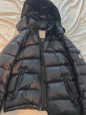 Mörkblå Moncler Maya small - Snygg svart dunjacka från Moncler med glansig finish, stor vadderad huva och dragkedja framtill. Jackan har två snedställda fickor med dragkedja och en ärmficka med Moncler-logga. Perfekt för kalla vinterdagar.