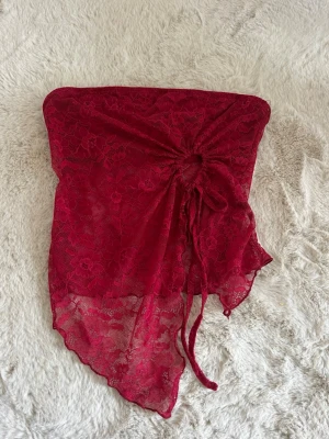 Vinröd spets tubtopp med snörning - Säljer en vinröd tubtopp i spets med asymmetrisk nederkant och snygg snörning framtill. Toppen är axelbandslös och har ett blommigt spetsmönster. Storlek XS, aldrig använd 
