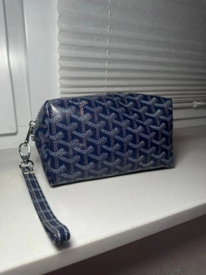 Blå necessär bag med mönster - Snygg blå necessär väska med klassiskt vitt och grått grafiskt mönster. Väskan har en avtagbar handledsrem och silverfärgade metalldetaljer. Perfekt för att organisera småsaker eller smink. Tillverkad i tåligt material med lyxig känsla.