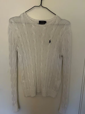 Vit kabelstickad tröja Polo Ralph Lauren - Kabelstickad vit tröja från Polo Ralph Lauren med rund halsringning och broderad logga på bröstet. Långärmad modell med ribbade muddar och nederkant. Klassisk och stilren design som passar till mycket.