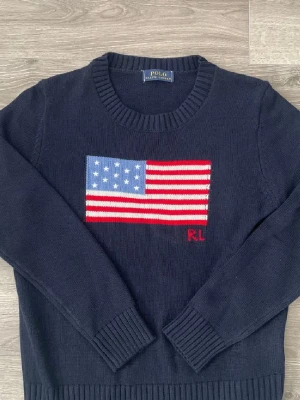 Mörkblå stickad tröja Polo Ralph Lauren - Mörkblå stickad tröja från Polo Ralph Lauren med amerikansk flagga i rött, vitt och blått på bröstet samt RL-broderi. Tröjan har rund halsringning och ribbstickade muddar vid ärmslut och nederkant. Klassisk och tidlös design. Blev ett mini hål i ena hörnat av flaggan men det är lagat. Hör av er vid frågor och skicka gärna prisförslag 😄✅