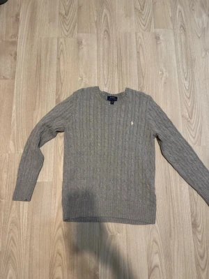 Grå kabelstickad tröja Polo Ralph Lauren - Snygg grå kabelstickad tröja från Polo Ralph Lauren med rund halsringning och diskret broderad logga på bröstet. Tröjan har långa ärmar och ribbade muddar vid ärmslut och nederkant. Perfekt för lager på lager och enkel att matcha. Tröjan har ett litet hål vid armhålan men syns inte