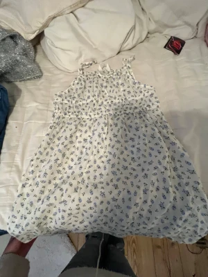 Vit blommig sommarklänning med rosett - Söt vit kortklänning med blått blommönster och smala axelband. Klänningen har en liten rosett framtill vid urringningen och är gjord i ett lätt, luftigt material som passar perfekt för varma dagar.