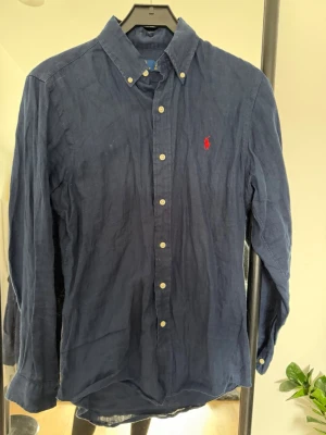 Marinblå linneskjorta från Ralph Lauren - Klassisk marinblå skjorta från Ralph Lauren i linne med knappar framtill och ikonisk röd logga broderad på bröstet. Skjortan har lång ärm och button-down krage, perfekt för en stilren och avslappnad look.