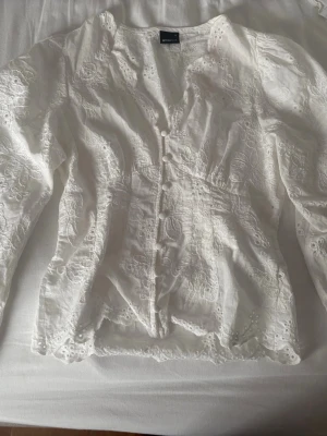 Vit broderad blus från Gina Tricot - Säljer en vit blus från Gina Tricot med vackra broderade detaljer och knappar framtill. Blusen har långärmade puffärmar och är gjord i ett lätt material med spetsmönster och dekorativa kanter. 