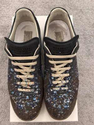 Maison Margiela gat strl44 - Unika sneakers från Maison Margiela med svart och brunt läder, dekorerade med vita, blå och orange färgstänk.Perfekt för dig som gillar statement-plagg och konstnärlig stil. Skorna är i stort sett omöjliga och få tag på idag då de senast tillverkades 2017 och just de här paret är i sjukt bra skick för att vara så gammal. De här är för dig som vill sticka ut och veta att ingen annan kommer ha på sig samma sko som dig.