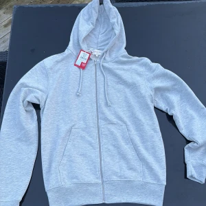 Ljusgrå Arsenal zip hoodie S - Ljusgrå hoodie från Arsenal F.C. med dragkedja framtill, broderad kanonlogga på bröstet och två fickor. Tröjan har huva med snörning. Ny pris 60 pund, köptes på Emirates Stadium. Passar M. Helt ny