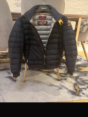 Svart dunjacka från Parajumpers - Snygg svart dunjacka från Parajumpers med quiltad design och gul detalj vid kragen. Jackan har dragkedja framtill, ribbade muddar och patch på ärmen. Perfekt för kalla vinterdagar och riktigt stilren look.