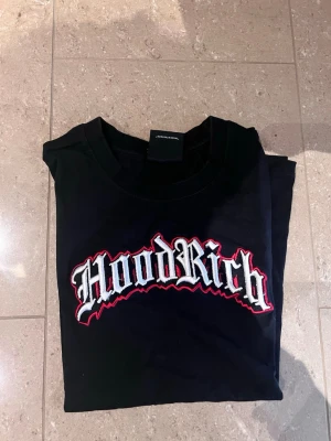 Svart Hoodrich t-shirt med tryck - Snygg svart t-shirt från Hoodrich med stort broderat logotyp-tryck i vitt och rött på bröstet. Klassisk rund hals och korta ärmar. Perfekt för dig som vill ha en streetwear-look med attityd.
