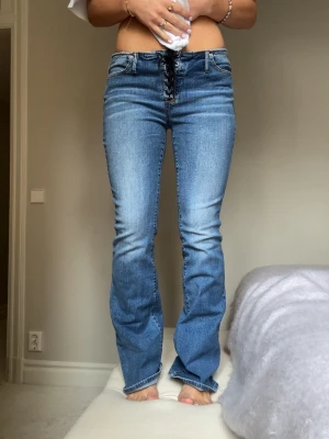 Blå bootcut jeans med snörning - Snygga blå bootcut jeans med unik snörning framtill istället för vanlig dragkedja. Jeansen har klassiska fickor både fram och bak och är tillverkade i stretchigt denimtyg för skön passform. Perfekta för dig som vill ha en cool och trendig look.