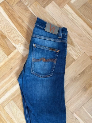 Blå jeans från Nudie Jeans - Snygga blå jeans från Nudie Jeans med klassisk femficksdesign och orangea sömmar. Jeansen har en rak passform och är tillverkade i denim med lätt slitning på framsidan och bakfickan. Perfekta för en avslappnad och trendig look.