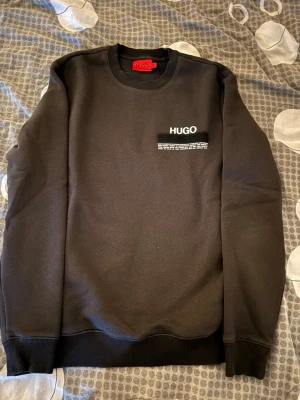 Svart sweatshirt från HUGO - Snygg svart sweatshirt från HUGO med rund halsringning och diskret logga på bröstet. Tröjan har ribbade muddar vid ärmslut och nederkant. Tillverkad i mjukt material som känns skönt mot huden. Perfekt för en clean och stilren look.