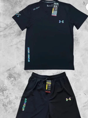 Svart träningsset från Under Armour - Snyggt svart träningsset från Under Armour med t-shirt och shorts. Båda plaggen har logga och text i turkos och vit. Tillverkade i ett lätt och snabbtorkande material som passar perfekt till träning. T-shirten har rund hals och shortsen har resår i midjan.