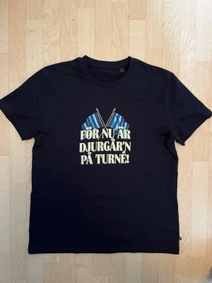 Djurgår'n t-shirt med tryck - Svart t-shirt med tryck fram och bak. Framsidan har texten 'FÖR NU ÄR DJURGÅR'N PÅ TURNÉ!' och två blå-randiga flaggor. Baksidan har en illustration av en supporter i blå-randig tröja med bengal och texten 'SE BENGALERNAS SKEN'. Klassisk passform.