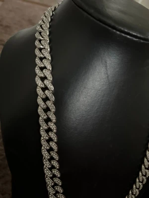Silverfärgat cuban chain halsband - Säljer ett chunky halsband i cuban chain-stil med silverfärgade länkar täckta av gnistrande stenar. Halsbandet har en modern och iögonfallande design som verkligen sticker ut. Perfekt för dig som gillar statement-smycken och vill addera lite extra bling till din look.