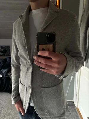 Kavaj cardigan  - Snygg ljusgrå kavaj med två stora framfickor och klassisk krage. Kavajen har en avslappnad passform och är tillverkad i ett mjukt, strukturerat tyg som ger en modern look. Perfekt att slänga över en t-shirt för en clean stil.