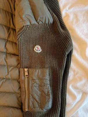 Moncler grön stickad cardiganjacka - Snygg grön cardiganjacka från Moncler med stickade ärmar och quiltad kropp. Jackan har huva, dragkedja framtill och fickor med dragkedja på ärmen. 