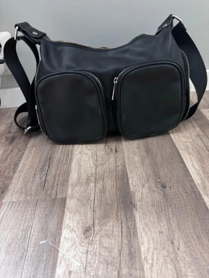 Svart axelväska med dubbla fickor - Snygg svart axelväska i slätt konstläder med två stora framfickor med dragkedja och justerbar axelrem i textil. Väskan har även en praktisk bakficka med dragkedja och silverfärgade detaljer. Perfekt för dig som gillar en stilren och funktionell look.