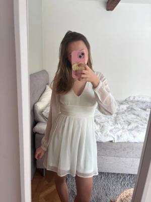 Studentklänning - Kort vit klänning med v-ringad hals och långa, transparenta ärmar. Klänningen har markerad midja och lätt utsvängd kjol. Materialet känns luftigt och flowy, perfekt för varma dagar.