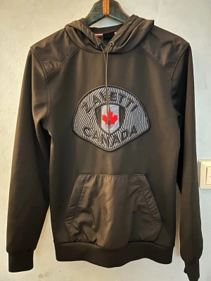 Svart hoodie från Zavetti Canada - Svart hoodie från Zavetti Canada med stor broderad logga och röd lönnlöv på bröstet. Klassisk känguruficka framtill, dragsko i huvan och ribbade muddar. Materialet är mjukt och slitstarkt, perfekt för en avslappnad streetwear-look. Först till kvarn som gäller!