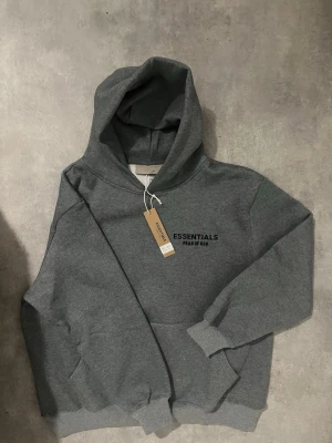 Grå Essentials Fear of God hoodie - Snygg grå hoodie från Essentials Fear of God med svart logga tryckt på bröstet och ryggen. Klassisk känguruficka framtill, ribbade muddar och en stor huva. Perfekt för dig som gillar streetwear och vill ha en stilren look. Jag och hoodien är size S men den var för stor för mig så jag rekommenderar den till någon som är size M eller någon som knappast passar i size S