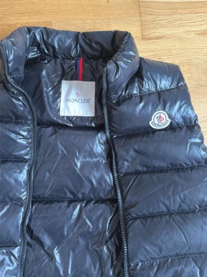 Svart dunväst från Moncler - Snygg svart dunväst från Moncler med hög krage och quiltad design. Västen har dragkedja framtill och Moncler-logga på bröstet. Tillverkad i glansigt material som ger en modern look. Perfekt att slänga över en hoodie eller tröja för extra värme.
