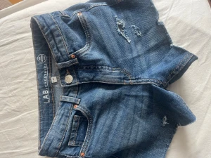 Blå denimshorts från LAB - Säljer ett par klassiska blå denimshorts från LAB med råa kanter och slitna detaljer. Shortsen har fem fickor, knappgylf och kontrastsömmar. Perfekta för varma dagar och en avslappnad stil.
